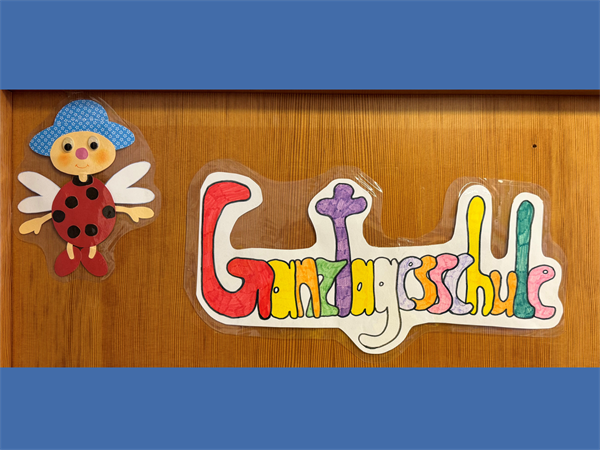 Schriftzug Ganztagesschule in bunten Farben und selbstgemalt; links daneben ein selbstgestalteter Marienkäfert mit Beinen und Händen und einem blauen Hut; oben und unten jeweils ein mittelblauer Balken